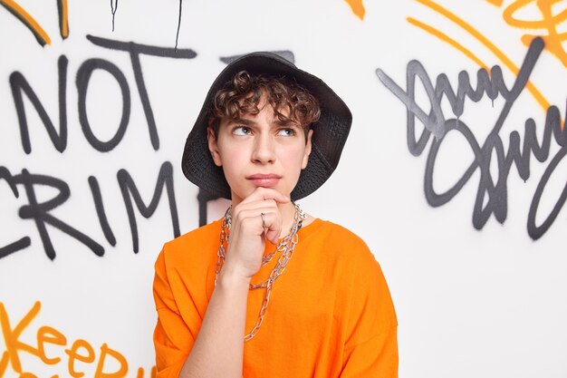Nachdenklicher Mann im Streetstyle-Outfit hält Kinn trägt schwarzen Hut und orange T-Shirt-Posen neben Graffiti-Wand hat nachdenklichen Ausdruck