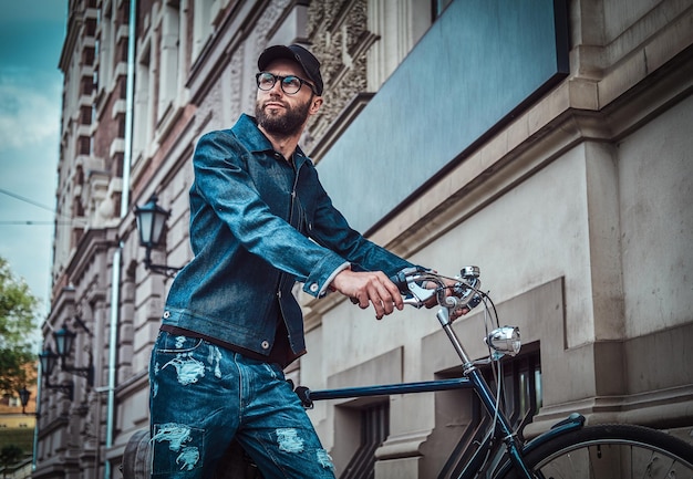 Nachdenklicher Mann in Brille, Denim und Mütze steht auf der Straße. Er hält sein Fahrrad. Der Mensch schaut weg. Gebäude und Himmel im Hintergrund.