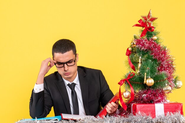 Nachdenklicher nachdenklicher junger Mann in einem Anzug, der am Tisch sitzt und Dokumente Weihnachtsbaum und Geschenke prüft