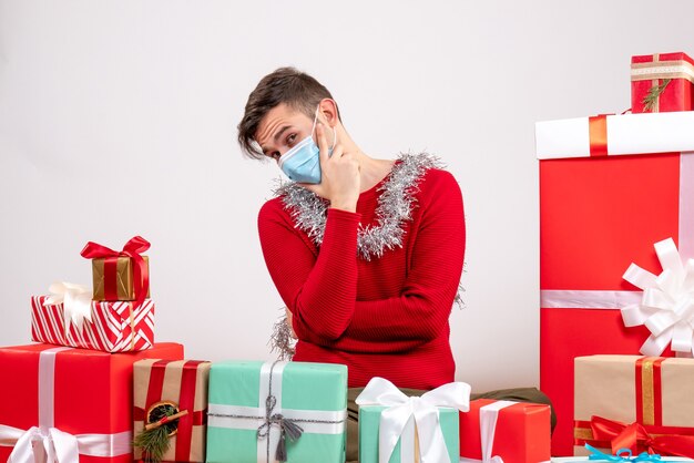 Nachdenklicher nachdenklicher junger Mann mit Maske, die nach hinten zeigt, um Weihnachtsgeschenke sitzend