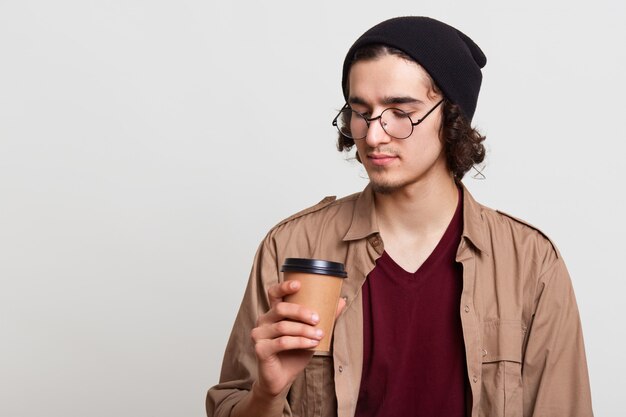 Nachdenklicher nachdenklicher Yougster, der eine Tasse Kaffee mit Papier hat, ein heißes Getränk in einer Hand hält, es aufmerksam betrachtet, isoliert auf hellgrau posiert und in der Pause ist. Jugendkonzept.