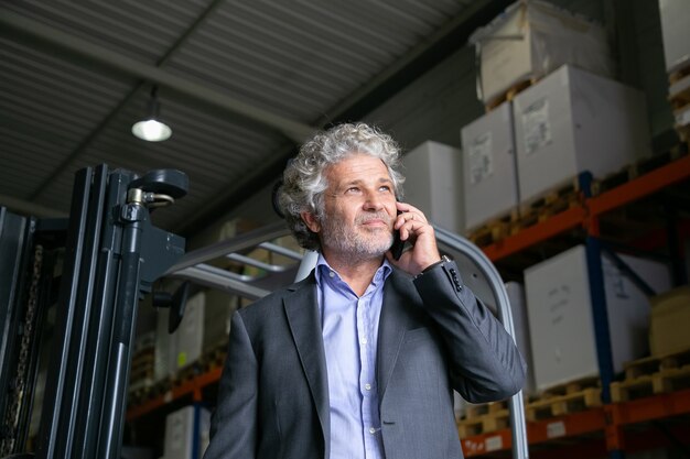 Nachdenklicher reifer Geschäftsmann, der nahe Gabelstapler im Lager steht und auf Handy spricht. Regale mit Waren im Hintergrund. Geschäfts- oder Logistikkonzept