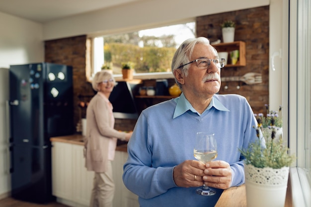 Nachdenklicher reifer Mann, der Wein trinkt und an etwas in der Küche denkt Seine Frau bereitet im Hintergrund Essen zu