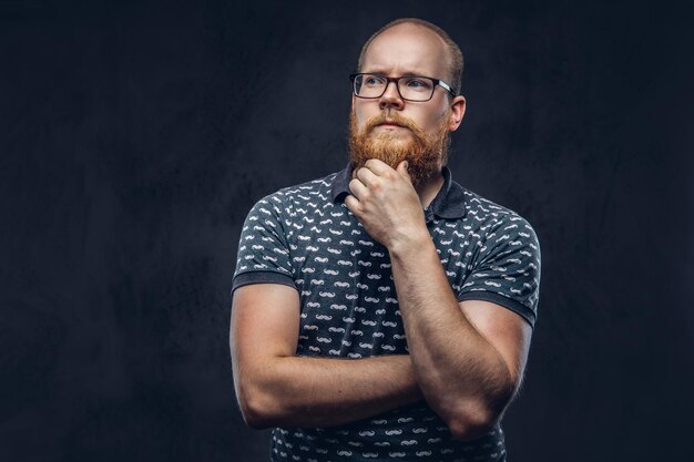 Nachdenklicher rothaariger bärtiger Mann mit Brille, gekleidet in ein T-Shirt, das mit einer Hand am Kinn posiert. Getrennt auf einem dunklen strukturierten Hintergrund.