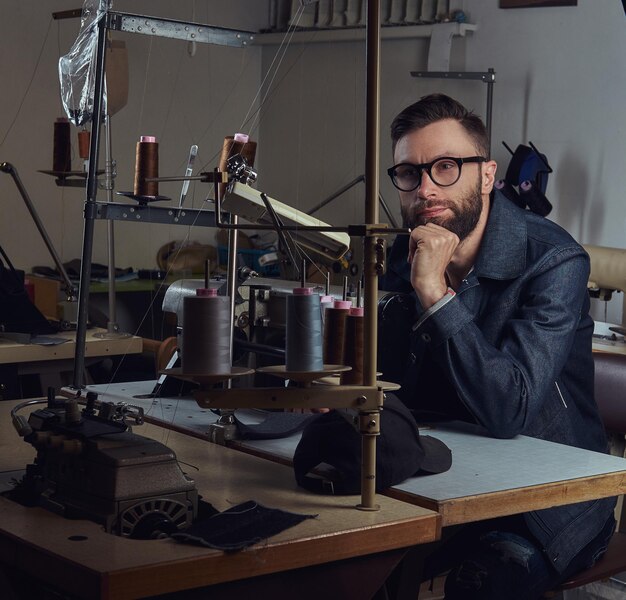 Nachdenklicher Schneider, der in einer Nähwerkstatt mit der Hand am Kinn am Tisch sitzt.