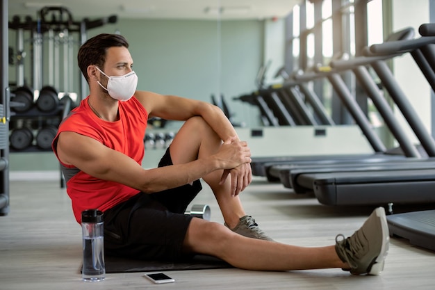 Nachdenklicher Sportler mit schützender Gesichtsmaske, der sich im Fitnessstudio auf dem Boden entspannt