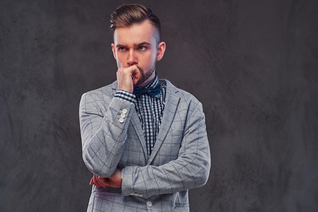 Nachdenklicher stilvoller bärtiger Mann mit Frisur in einem eleganten grauen Retro-Anzug und Fliege, der in einem Studio posiert. Isoliert auf grauem Hintergrund.