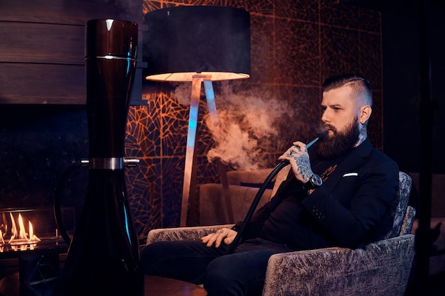 Nachdenklicher tätowierter Mann sitzt auf dem Sessel und fängt an, Wasserpfeife zu rauchen. Es gibt schönen Dampf.