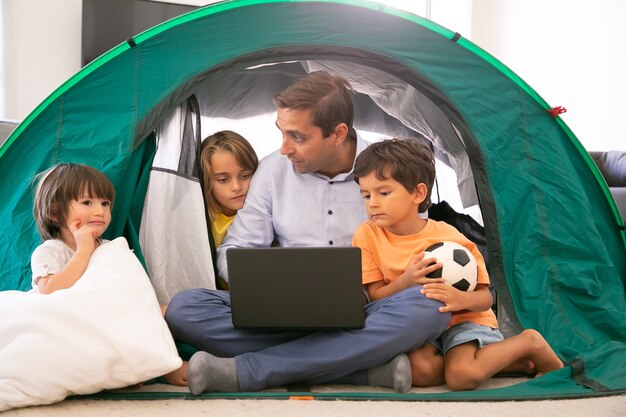 Nachdenklicher Vater, der mit gekreuzten Beinen mit Kindern im Zelt zu Hause sitzt und Laptop hält. Nette Kinder, die Film auf tragbarem Computer mit kaukasischem Vater ansehen. Kindheits-, Familienzeit- und Wochenendkonzept