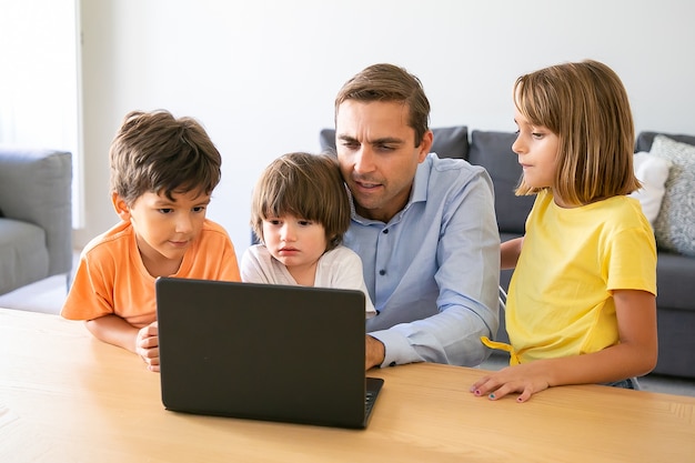 Nachdenklicher Vater und Kinder beobachten etwas auf Laptop-Bildschirm. Fokussierter kaukasischer Vater sitzt am Tisch, umgeben von entzückenden Kindern. Konzept für Kindheit, Familie, Vaterschaft und digitale Technologie