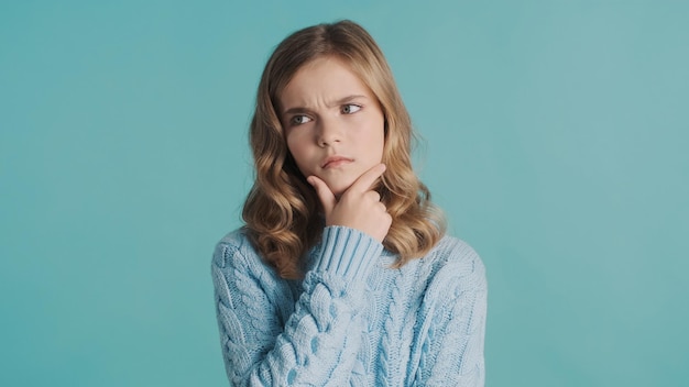 Nachdenkliches blondes Teenager-Mädchen, das sich Sorgen macht und die Hand am Kinn hält, über etwas nachzudenken, über blauem Hintergrund Gesichtsausdruck