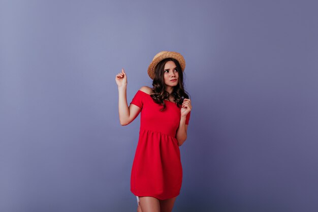 Nachdenkliches gut gekleidetes Mädchen, das auf lila Wand aufwirft. Attraktive dunkelhaarige Frau im roten Kleid und im Strohhut, die Fotoshooting genießt.