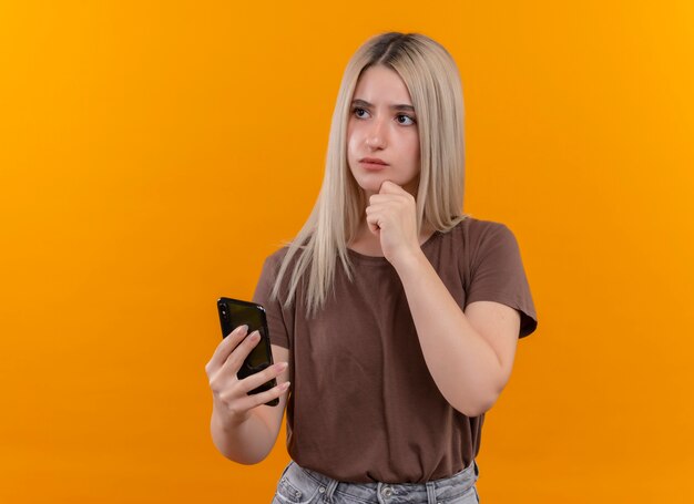 Nachdenkliches junges blondes Mädchen, das Handy mit Hand auf Kinn auf isolierter orange Wand mit Kopienraum hält