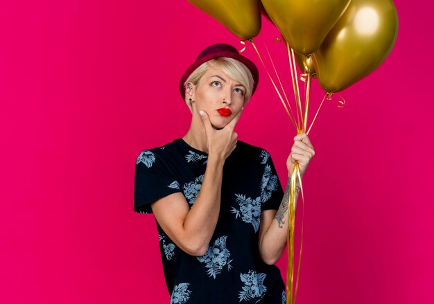 Nachdenkliches junges blondes Partygirl, das Partyhut hält, der Ballons hält, die Kinn berühren, lokalisiert auf purpurrotem Hintergrund mit Kopienraum
