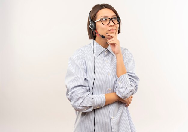 Nachdenkliches junges Callcenter-Mädchen, das Brille und Headset trägt und Seite betrachtet, die Hand auf Kinn lokalisiert auf weißer Wand setzt