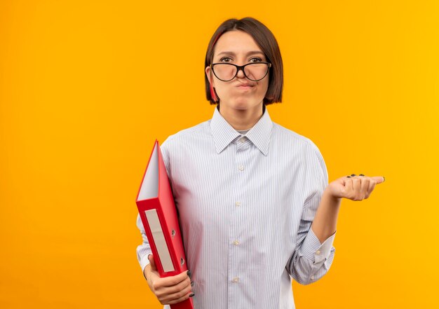 Nachdenkliches junges Callcenter-Mädchen, das eine Brille hält, die Ordner zeigt, der auf Seite lokalisiert auf orange Wand zeigt