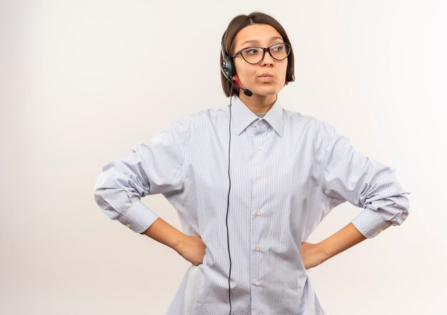 Nachdenkliches junges Callcenter-Mädchen, das eine Brille und ein Headset trägt, die Hände auf Taille setzen, die Seite lokalisiert auf weißer Wand betrachten