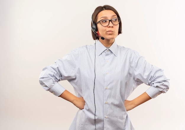 Nachdenkliches junges Callcenter-Mädchen, das eine Brille und ein Headset trägt, die Hände auf Taille setzen, die Seite lokalisiert auf weißer Wand betrachten