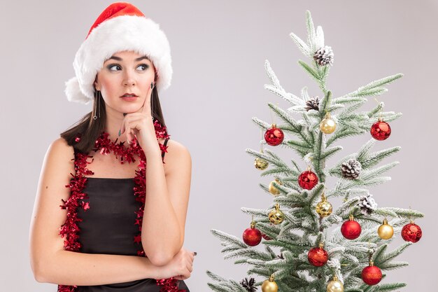 Nachdenkliches junges hübsches kaukasisches Mädchen, das Weihnachtsmütze und Lametta-Girlande um den Hals trägt, der nahe verziertem Weihnachtsbaum steht und Hand auf Kinn hält, das Seite lokalisiert auf weißer Wand betrachtet