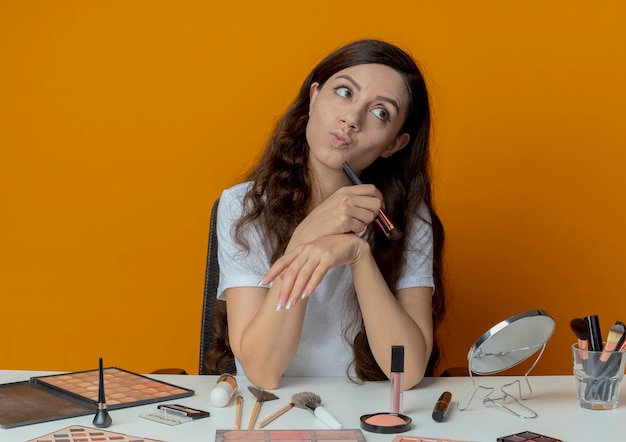 Nachdenkliches junges hübsches Mädchen, das am Make-up-Tisch mit Make-up-Werkzeugen sitzt, errötenden Pinsel hält und Kinn damit berührt, das Seite betrachtet, die auf orange Hintergrund lokalisiert wird