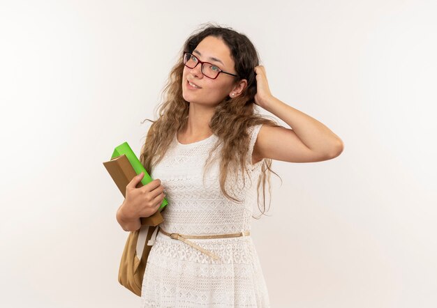 Nachdenkliches junges hübsches Schulmädchen, das Brille und Rückentasche hält Bücher hält, die Hand auf Kopf setzen und Seite lokalisiert auf weißer Wand betrachten