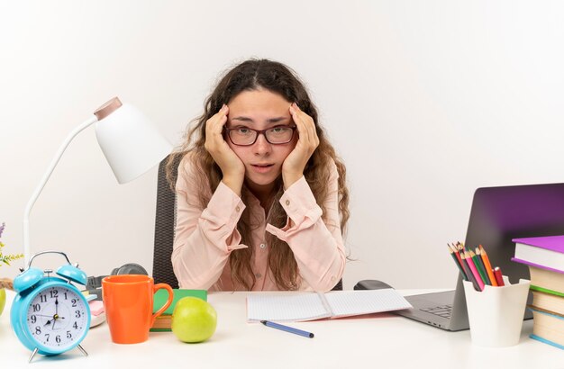 Nachdenkliches junges hübsches Schulmädchen, das eine Brille trägt, die am Schreibtisch mit Schulwerkzeugen sitzt, die ihre Hausaufgaben machen, die ihren Kopf lokalisiert auf weißer Wand halten