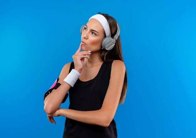 Nachdenkliches junges hübsches sportliches Mädchen, das Stirnband und Armband und Kopfhörer mit Telefonarmband trägt, die Seite betrachten und Hand auf Kinn lokalisiert auf blauem Raum setzen