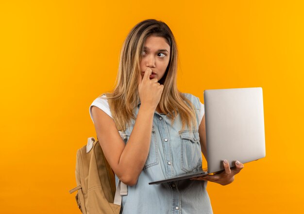 Nachdenkliches junges hübsches Studentenmädchen, das Rückentasche hält Laptop hält, das Seite mit Finger auf Lippen lokalisiert auf orange Wand betrachtet