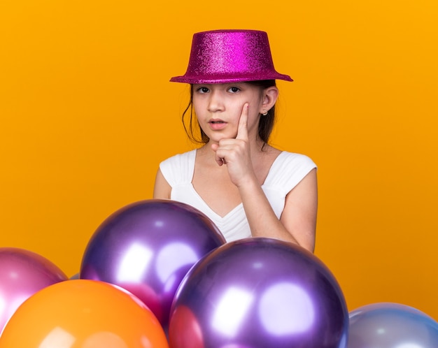 Nachdenkliches junges kaukasisches mädchen mit lila partyhut, das die hand auf das kinn legt und mit heliumballons isoliert auf orangefarbener wand mit kopierraum steht