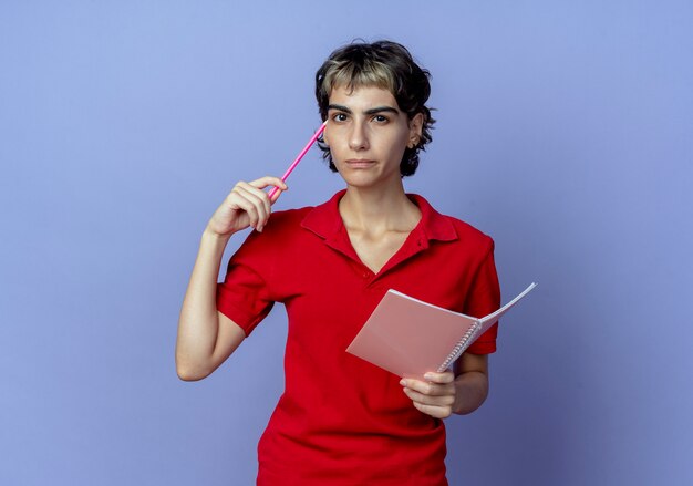 Nachdenkliches junges kaukasisches Mädchen mit Pixie-Haarschnitt, der Stift und Notizblock berührt Gesicht mit Stift lokalisiert auf lila Hintergrund mit Kopienraum