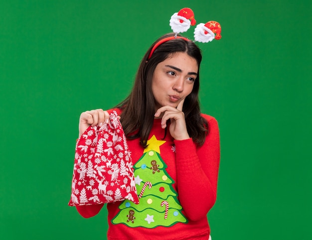 Nachdenkliches junges kaukasisches Mädchen mit Weihnachtsmann-Stirnband legt Finger auf Kinn und hält Weihnachtsgeschenktüte, die Seite lokalisiert auf grünem Hintergrund mit Kopienraum betrachtet