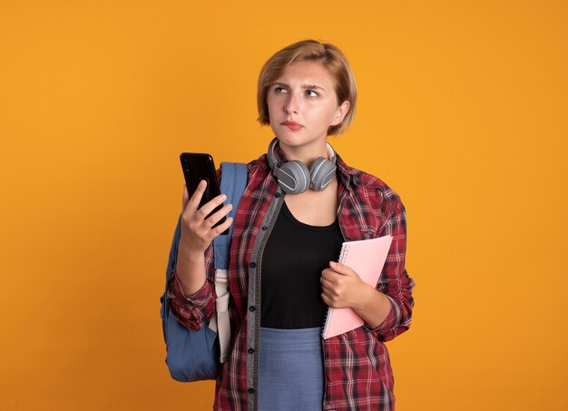 Nachdenkliches junges slawisches Studentenmädchen mit Kopfhörern, das Rucksack trägt, hält Notebook und Telefon, das auf die Seite schaut