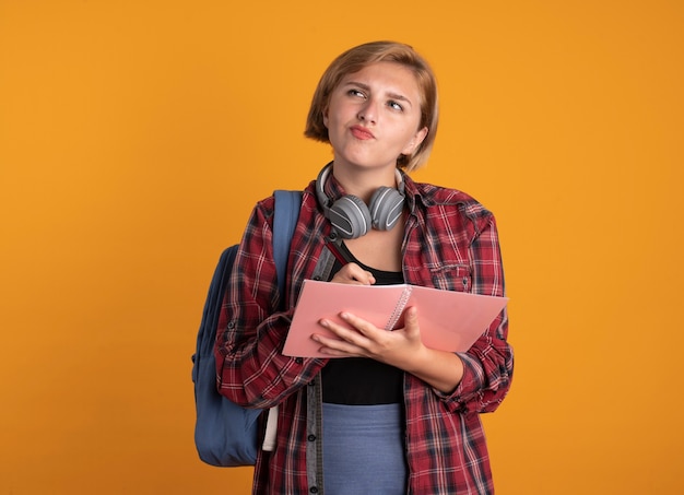 Nachdenkliches junges slawisches Studentenmädchen mit Kopfhörern, das Rucksack trägt, hält Stift und Notizbuch auf der Seite