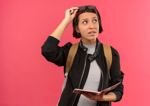 Nachdenkliches junges Studentenmädchen, das Brille auf Kopf- und Rückentasche hält, die Notizblock und Brille hält, die Seite lokalisiert auf rosa Wand betrachten