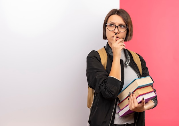 Nachdenkliches junges Studentenmädchen, das Brille und Rückentasche hält Bücher hält, die vor der weißen Wand stehen, die ihr Kinn berührt, die Seite betrachtet, die auf rosa Wand lokalisiert wird