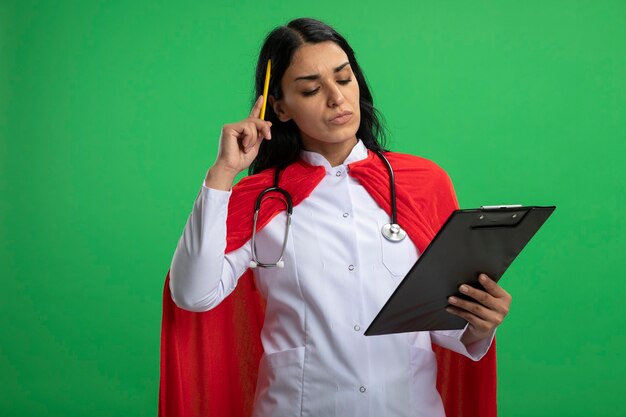 nachdenkliches junges Superheldenmädchen, das medizinisches Gewand mit Stethoskop hält und Klemmbrett hält, das Stift auf Kopf lokalisiert auf Grün setzt