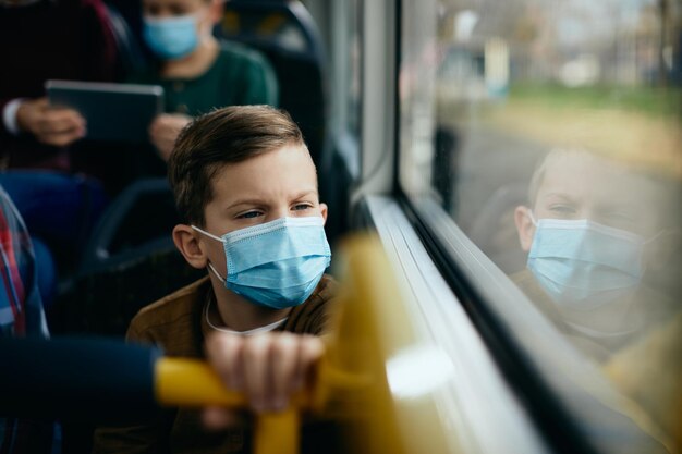 Nachdenkliches Kind mit Gesichtsmaske, das während der Busfahrt durch das Fenster schaut