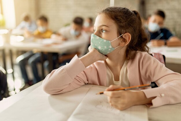 Nachdenkliches Mädchen, das eine schützende Gesichtsmaske trägt, während es im Unterricht im Notizbuch schreibt