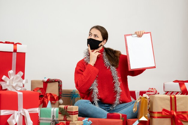 nachdenkliches Mädchen mit schwarzer Maske, die Dokumente hält, die um Geschenke auf Weiß sitzen