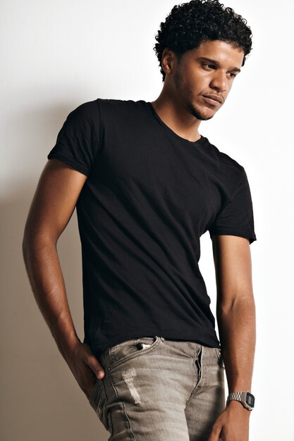 Nachdenkliches muskulöses schwarzes junges Model in einem schlichten schwarzen Baumwoll-T-Shirt und Jeans mit der rechten Hand in der hinteren Jeanstasche an der weißen Wand.