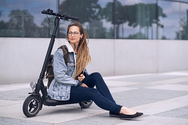 Nachdenkliches nettes Mädchen mit Brille und Tätowierungen an Hals und Händen chillt auf ihrem eigenen Roller.