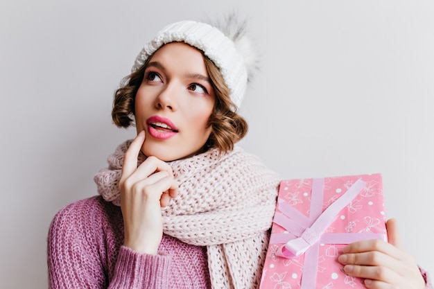 Nachdenkliches spektakuläres Mädchen im niedlichen Hut, der mit rosa Geschenkbox aufwirft. ekstatische Frau trägt einen gestrickten Schal, der an etwas denkt und Neujahrsgeschenk hält.