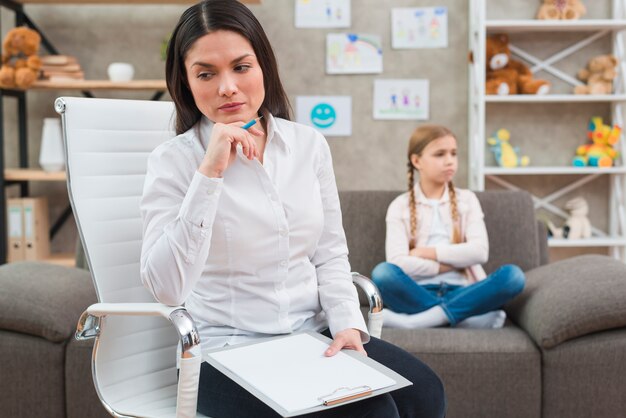 Nachgedachtes Psychologeholding färbte den blauen Bleistift und Klemmbrett, die vor dem traurigen Mädchen sitzen auf Sofa sitzen