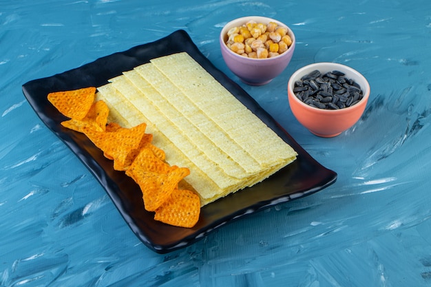 Nachos-Chips und lange Chips auf einer Platte neben Samen und Kichererbsen, auf der blauen Oberfläche.