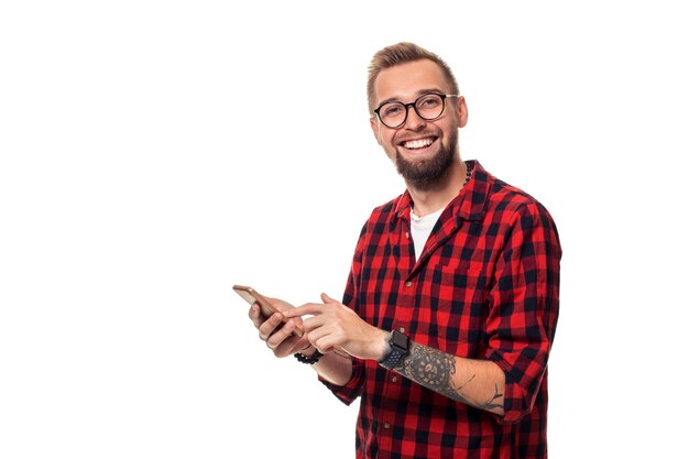 Nachricht an Freund. Hübscher junger Mann, der Telefon mit Lächeln beim Stehen gegen weißen Hintergrund verwendet. Studioaufnahme. Junger Hipster-Mann in kariertem Hemd und Brille