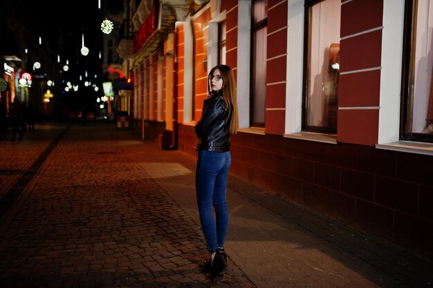 Nachtporträt eines Mädchenmodells mit Brille, Jeans und Lederjacke mit Rucksack auf den Straßen der Stadt