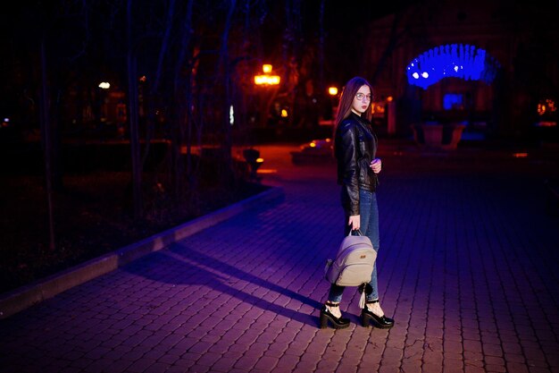 Nachtporträt eines Mädchenmodells mit Brille, Jeans und Lederjacke mit Rucksack in den Händen gegen Blaulichtgirlande der Stadtstraße