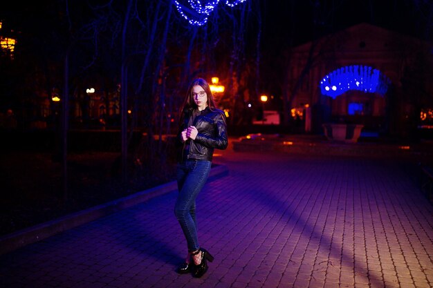 Nachtporträt von Mädchenmodellabnutzung auf Brillenjeans und Lederjacke gegen Blaulichtgirlande der Stadtstraße