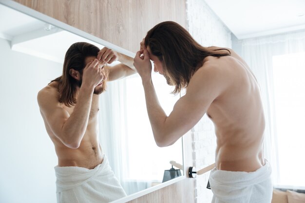 Nackter bärtiger junger Mann im Handtuch, der Pickel nahe Spiegel drückt