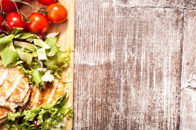 Nahaufnahme auf gegrillter Hähnchenbrust auf frisch geschnittenem Salat neben einem Bündel Tomaten auf einem Holzbrett